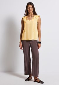 Top sans manches jaune avec un col en V et des cordons de serrage, agrémenté d'accents de volants texturés aux épaules ; porté avec un pantalon brun ample et des sandales noires.