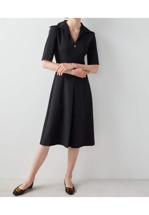 Vrouw in een zwart knielang jurkje met korte mouwen en kraag, zwarte platte schoenen met bruine strikjes, staand tegen een witte muur.