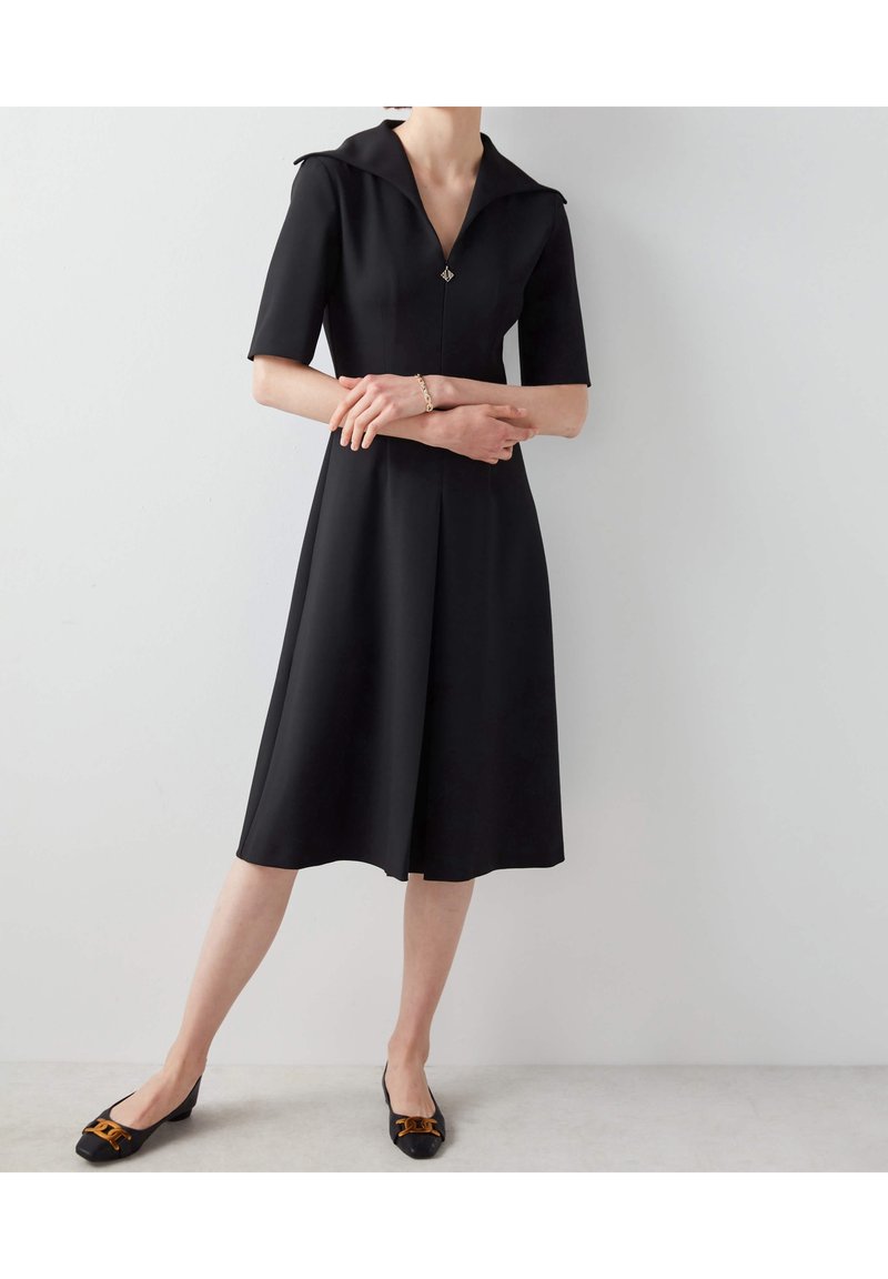 Femme en robe noire mi-longue à manches courtes et col, portant des ballerines noires avec des nœuds marron, debout contre un mur blanc.