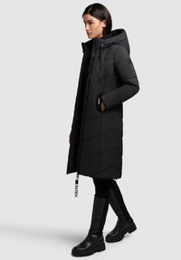 Cappotto lungo imbottito nero con cappuccio e zip, caratterizzato da un motivo a chevron e dettagli neri. Indossato con stivali neri fino al ginocchio.