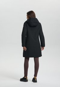 Manteau imperméable noir avec capuche, ornée d'un logo texturé sur le col et de longues manches. Porté avec des leggings marron et des baskets noires.