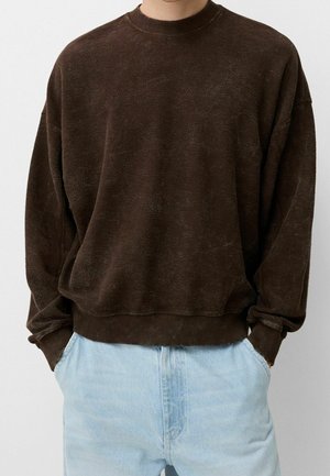 Person trägt einen locker sitzenden dunkelbraunen Pullover mit Rundhalsausschnitt, gerippten Bündchen und Saum, kombiniert mit hellblauen Jeans, die Hände in den Taschen.