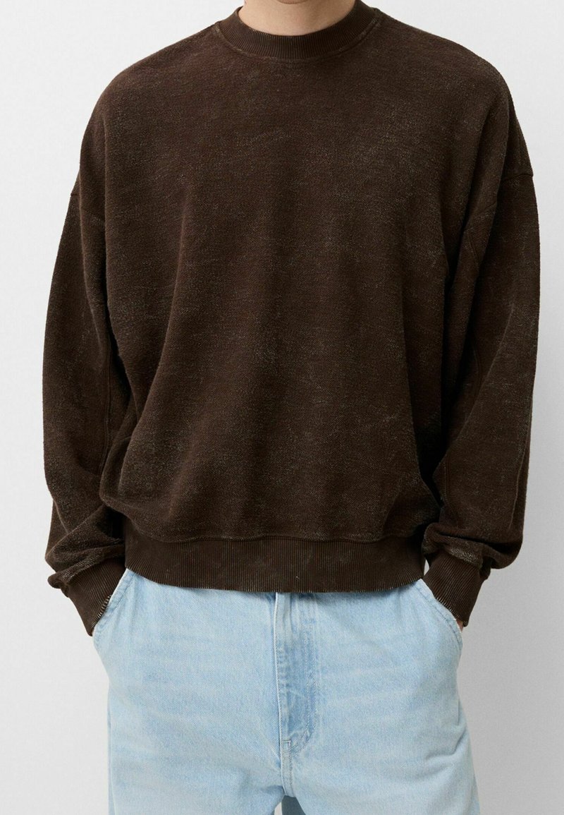 Personne portant un pull à col rond marron foncé ample avec des poignets et un ourlet côtelés, associé à un jean bleu clair, mains dans les poches.
