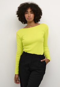 Dlouhý rukáv v jasné neonově žluté, vyrobený z žebrované látky, s off-shoulder výstřihem, kombinovaný s černými kalhotami s vysokým pasem.