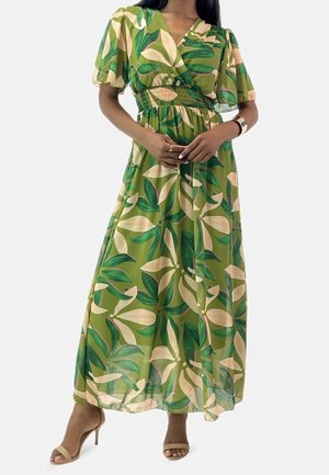 Donna che indossa un vestito lungo verde con stampa di foglie beige e verde scuro, maniche a sbuffo e sandali beige con tacco alto.