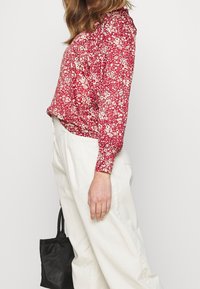 Rote Blumenbluse mit langen Ärmeln und gerafftem Ausschnitt. Hergestellt aus leichtem Stoff, mit kleinen weißen und rosa Blumen, kombiniert mit weißen Hosen.