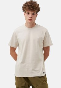 Camiseta de algodón beige con mangas cortas. Presenta un sutil logo blanco en el pecho y una etiqueta en el dobladillo. Textura suave y corte regular.