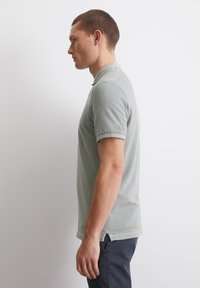 Młody mężczyzna stojący w profilu, ubrany w jasnoszary, krótko rękawowy polo i ciemne spodnie, na tle gładkiego białego tła.