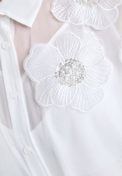 Hvid bluse med gennemsigtig stof, broderede blomsterapplikationer og pailleter, herunder to store blomster på skuldrene.