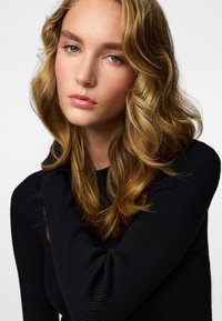 Svart ribbstickad tröja med hög hals, som har en åtsittande passform och långa ärmar, bärs av en modell med vågigt blont hår och naturlig makeup.