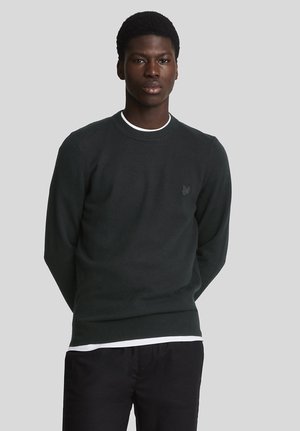 Mand iført sort rundhalset sweater over hvid skjorte og sorte bukser, stående med hænderne bag ryggen mod grå baggrund.