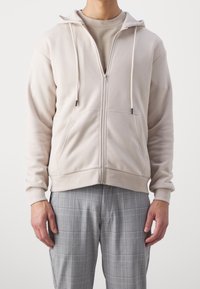 Felpa zip in beige chiaro con cappuccio, lacci e tasche laterali, abbinata a pantaloni a quadri grigi. Tessuto morbido, vestibilità rilassata.