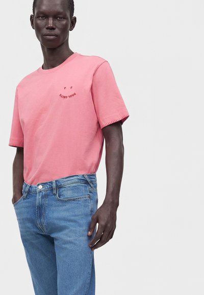 Modèle masculin portant un T-shirt rose à manches courtes avec un petit texte sur la poitrine et un jean bleu, debout devant un fond clair uni.
