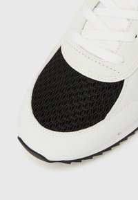 Valged tossud, millel on musta võrkmaterjaliga varbakate ja siledad ülekatteosad, rõhutatud valgete paelte ja tekstureeritud musta tallaga.