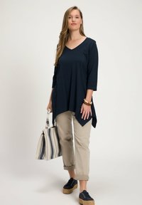 Top blu navy con scollatura a V e maniche a tre quarti, abbinato a pantaloni beige chiaro arrotolati e espadrillas blu navy, mentre tiene una borsa a tracolla a righe.