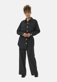 Donna con capelli ricci che indossa una camicia nera con bottoni dorati, pantaloni larghi abbinati e scarpe nere, in piedi con sicurezza.