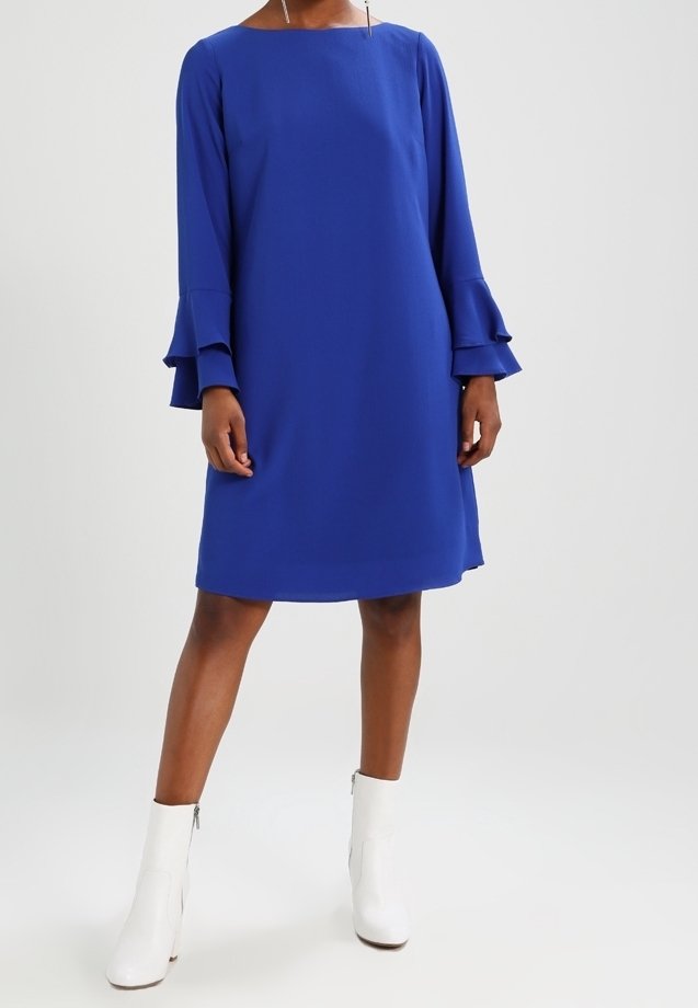 Femme portant une robe bleue longue jusqu'aux genoux, à manches longues avec des poignets cloche superposés, et des bottines blanches, sur un fond uni.