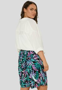 Valkoinen pitkähihainen paita, jossa on käärityt hihat, yhdistetty mustiin shortsit, joissa on värikäs trooppinen lehtikuosi vaaleanpunaisena, vihreänä ja sinisenä.