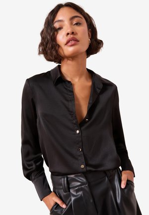 Donna con capelli corti e ricci che indossa una camicia nera in raso con bottoni e pantaloni in pelle neri, mani nelle tasche, espressione neutra.
