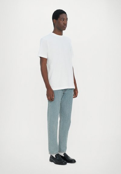 T-shirt en coton blanc, coupe décontractée, associé à un jean en denim bleu clair. Le modèle porte des chaussures noires à enfiler. Design simple et minimaliste.