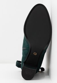 Bottine en daim vert foncé inclinée pour montrer la semelle noire lisse avec une bande antidérapante au talon et une fermeture éclair latérale.