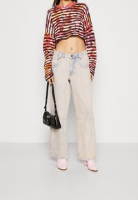 Nainen, jolla on monivärinen raidoitettu lyhyt neuletakki, vaaleat leveälahkeiset farkut, vaaleanpunaiset saappaat ja pieni musta käsilaukku.