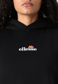 Čierna mikina s kapucňou vyrobená z mäkkej tkaniny, s predným logom "ellesse" v bielej farbe a farebným oblúkom nad ním. Kapucňa má voľný strih.