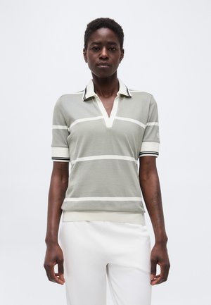 Persona che indossa una polo grigia a maniche corte con strisce orizzontali bianche e pantaloni bianchi, in piedi davanti a uno sfondo bianco semplice.