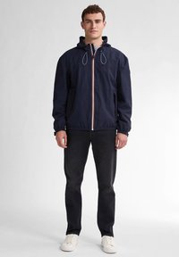 Marineblauwe windjack met een ritssluiting aan de voorkant, elastische manchetten en een capuchon met trekkoord. Voorzien van een meerkleurige streep in het midden. Zwarte jeans en witte sneakers.