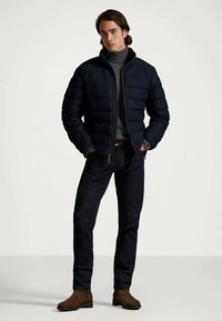 Nuori mies seisoo kädet taskuissa, päällään musta untuvatakki, harmaa poolopaita, tummat farkut ja ruskeat saappaat.