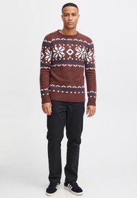 Bruine gebreide trui met witte sneeuwvlokpatronen, geribbelde manchetten, ronde halslijn, gecombineerd met zwarte broek en zwarte sneakers.
