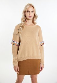 Beige gebreide trui met textuurstof, featuring kleurrijke franjes aan de mouwen, gecombineerd met een leren rok in bruin suede.