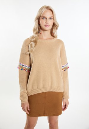 Beżowy sweter z dzianiny o teksturowanej tkaninie, z kolorowymi frędzlami na rękawach, zestawiony z brązową zamszową spódnicą.