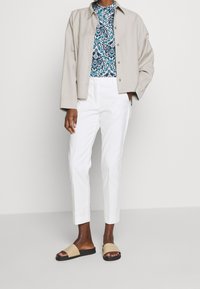 Osoba w beżowej zapinanej kurtce, niebieskiej bluzce w kwiatowy wzór, białych spodniach i beżowych klapkach stoi na tle jednolitego tła.