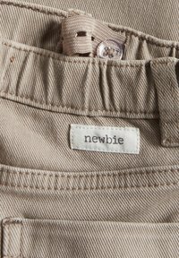 Beige denim-kangas, jossa näkyvät ompeleet, tasku ja "newbie"-merkki. Sisältää napin ja kudotun hihnan taskussa.