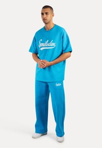 Camiseta de algodón azul con logo blanco de "Smilodox", combinada con pantalones de chándal azules a juego. Corte holgado, escote redondo y textura suave.