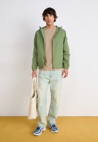 Light green zip-up jacket with elastic cuffs, beige t-shirt, faded blue jeans, and blue sneakers. Holding a cream tote bag.