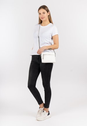 Bílá tričko, černé úzké džíny a bílé tenisky. Modelka drží malou bílou tašku přes rameno s kapsou na zip.