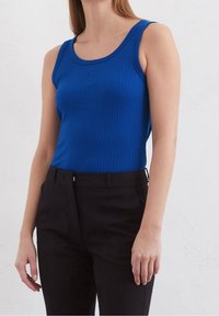 Top sin mangas ajustado y acanalado en un vibrante tejido azul, con escote redondeado y logo sutil en relieve. Combinado con pantalones negros.
