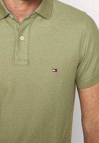 Homme portant un polo olive à manches courtes avec un petit logo rectangulaire rouge, blanc et bleu marine sur la poitrine gauche.
