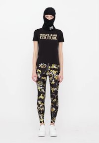Juodas marškinėliai su auksiniu užrašu "VERSACE JEANS COUTURE", derinami su juodomis leginsomis, turinčiomis spalvingą gėlių raštą, ir baltomis sportinėmis batelėmis.