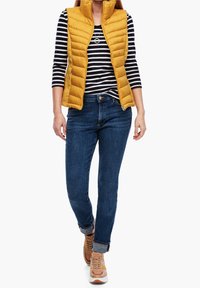 Żółta pikowana kamizelka na czarno-białym pasowanym topie, w połączeniu z niebieskimi dżinsami i brązowo-pomarańczowymi sneakersami. Zrolowane mankiety na dżinsach.