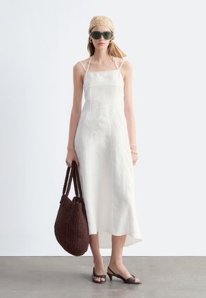 Femme portant une robe midi blanche sans manches, un foulard beige, des lunettes de soleil foncées, un sac cabas tressé marron et des talons ouverts foncés, debout sur un sol gris.