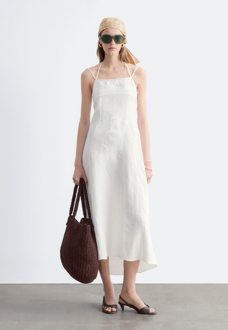 Femme portant une robe midi blanche sans manches, un foulard beige, des lunettes de soleil foncées, un sac cabas tressé marron et des talons ouverts foncés, debout sur un sol gris.