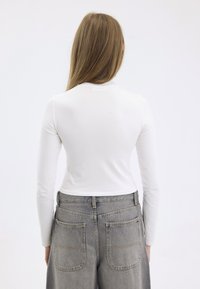 Langermet, tettsittende hvit topp laget av et glatt stoff, kombinert med høyt liv grå denim jeans som har store lommer og synlig søm.