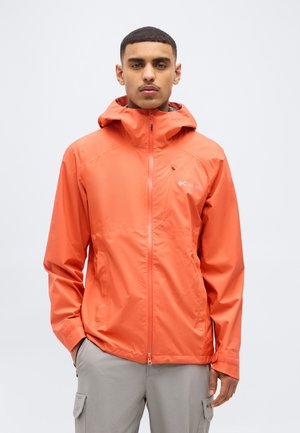 Giovane uomo che indossa una giacca antipioggia con cappuccio di un arancione intenso e pantaloni cargo grigio chiaro, in piedi su uno sfondo bianco semplice.