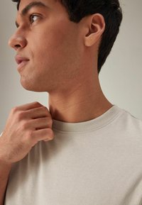 Tricou din bumbac de culoare bej deschis, cu decolteu rotund. Textură netedă, mâneci scurte, croială slim. Mână care apucă decolteul, concentrându-se pe țesătură.