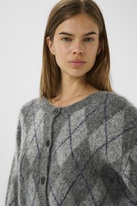 Young woman with straight brown hair wearing a gray fuzzy argyle-patterned cardigan with black buttons against a plain background.