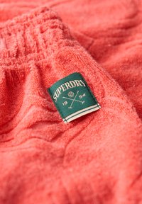 Un vêtement en tissu éponge corail avec une taille élastique, présentant une étiquette carrée verte avec le texte "Superdry" et un graphique de bâtons croisés.