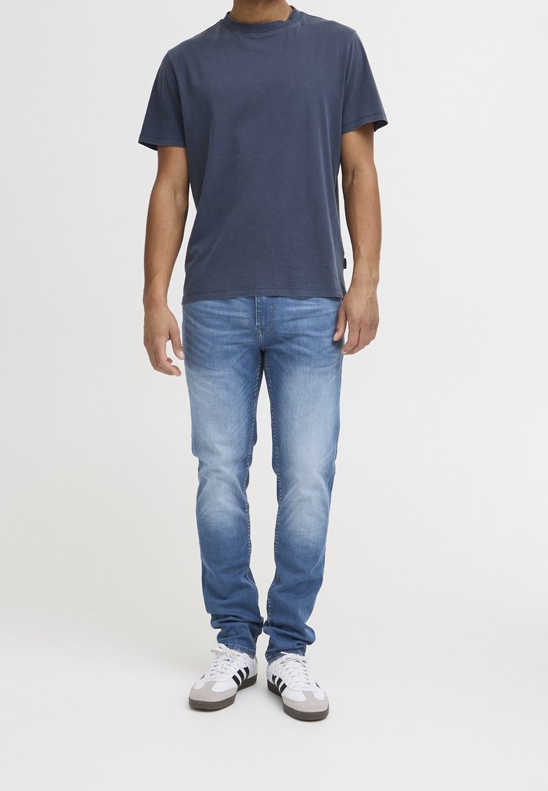 Homme portant un t-shirt bleu marine à manches courtes, un jean bleu clair et des baskets blanches avec des rayures noires, debout devant un fond uni.